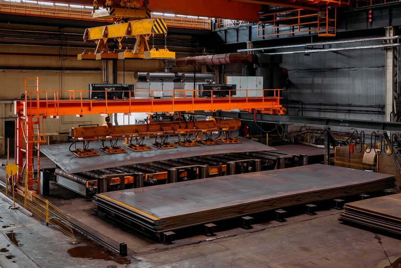 Planta industrial con grúas amarillas, maquinaria y grandes planchas de metal apiladas en un área de trabajo.