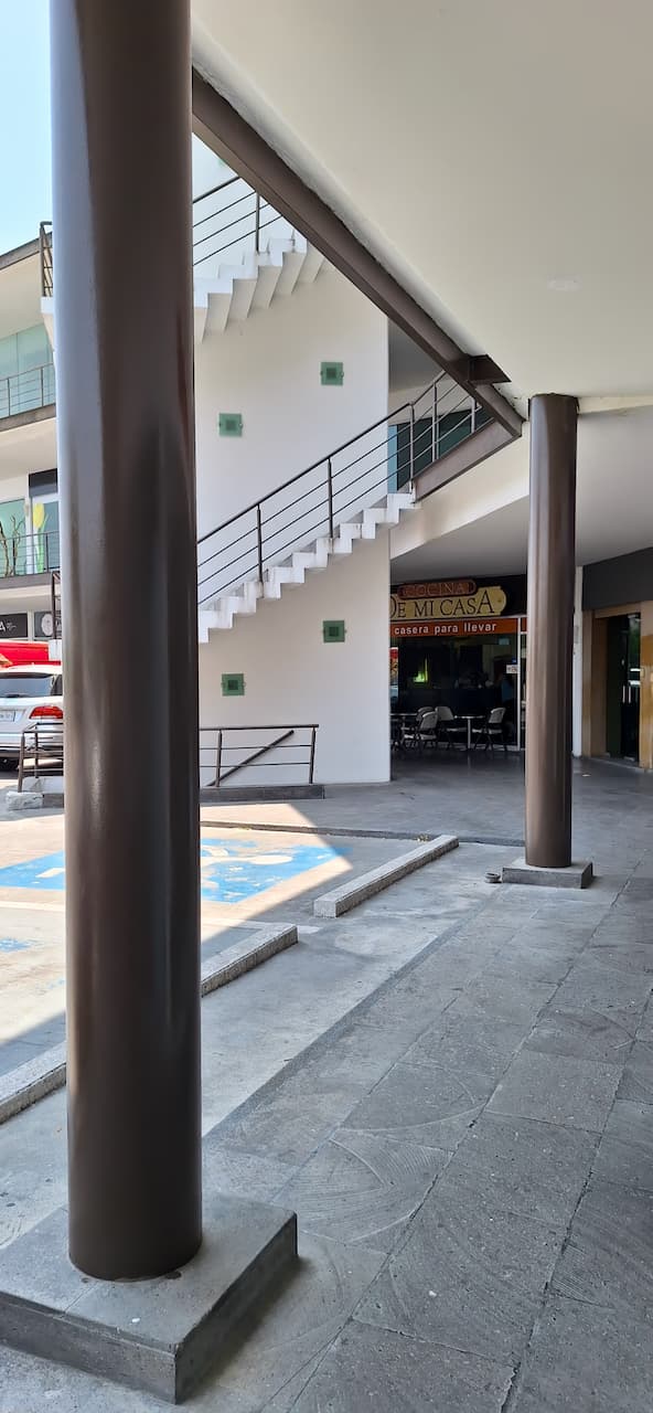 Espacio arquitectónico con escaleras blancas y columnas marrones, donde se observa un área comercial con mesas y sillas.