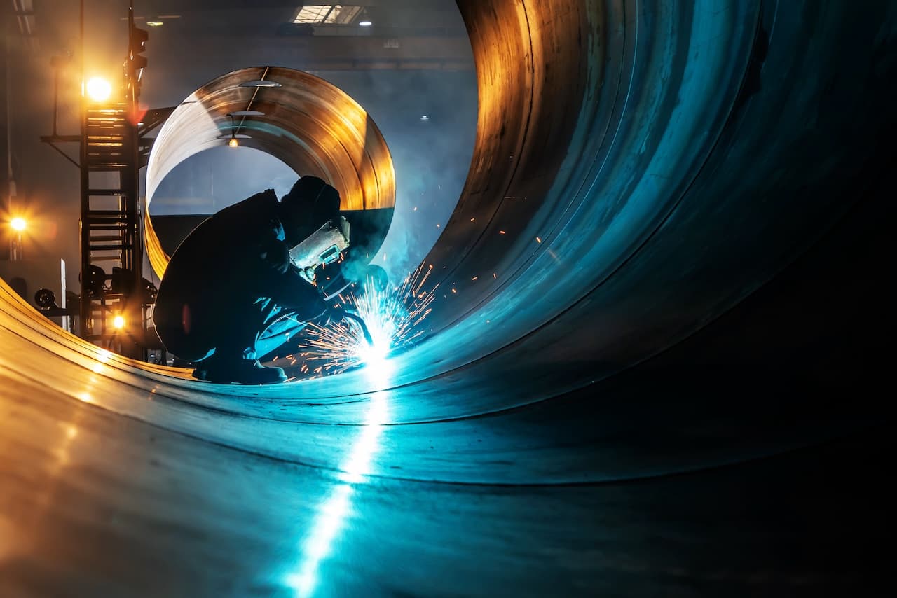 Soldador trabajando en la unión de tuberías metálicas con chispas y luz en un ambiente industrial.