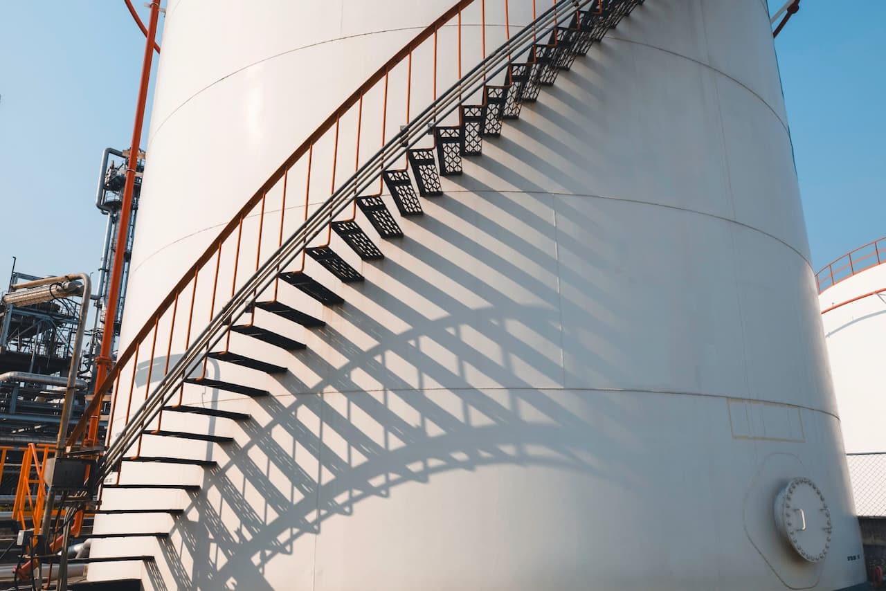 Escalera metálica negra en espiral proyectando sombras sobre un tanque blanco, con una estructura industrial al fondo y cielo despejado.