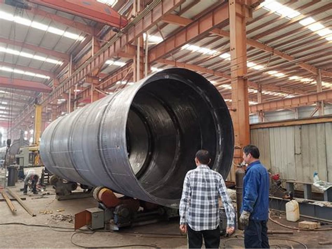 Tubería de acero de gran tamaño en una fábrica, con trabajadores observando y equipo industrial de fondo.