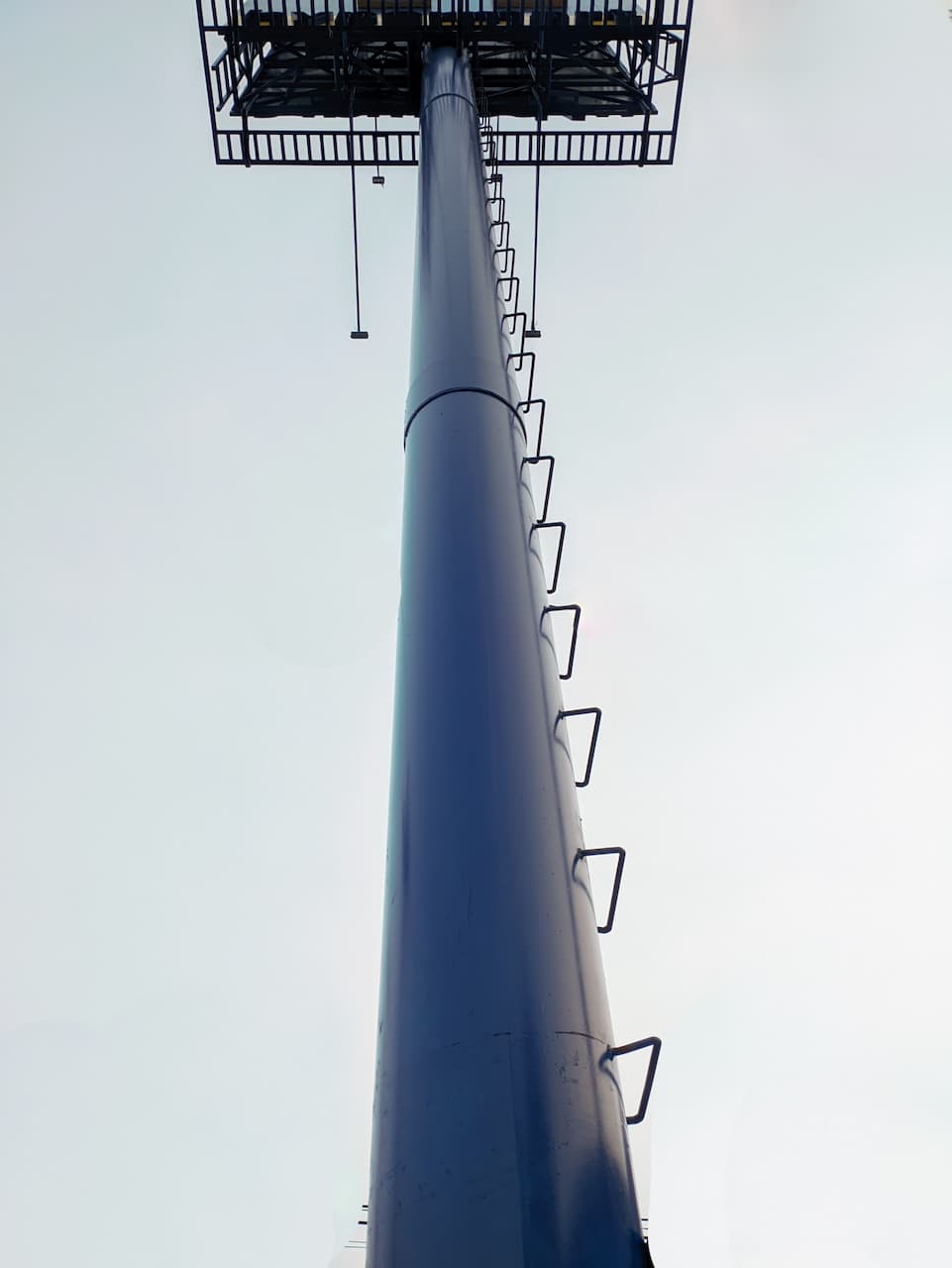 Torre vertical de metal con escaleras y señalización en la parte superior, contra un fondo de cielo claro.